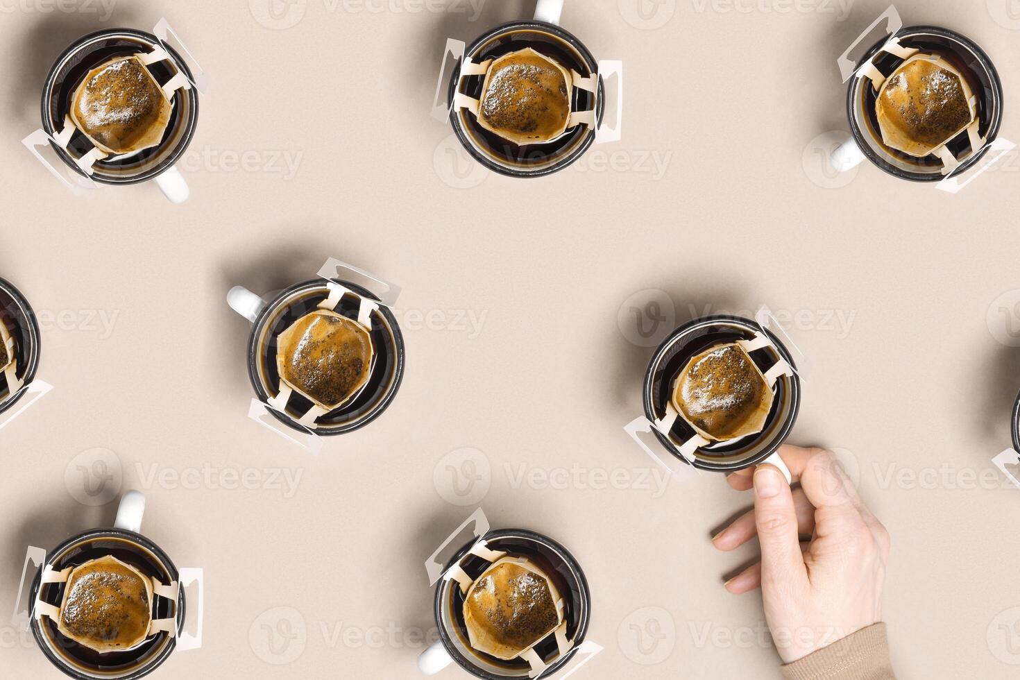 Pattern with black coffee cups, drip coffee bag with ground coffee for brewing in cup, paper bag drip coffee filter, female hand hold cup with drink, beige background, top view photo