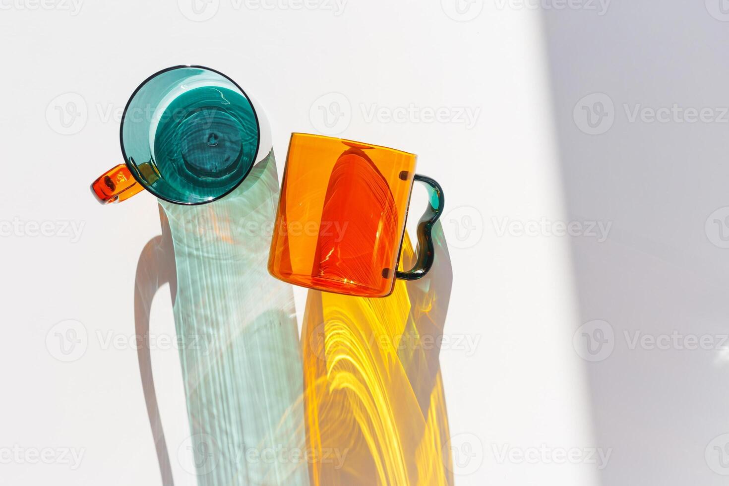 Orange and green Glass Mugs with bright sunlight Shadows, transparent cups casting dynamic, vibrant lights reflections and shadows on light white, creative aesthetic top view, minimal style, closeup photo