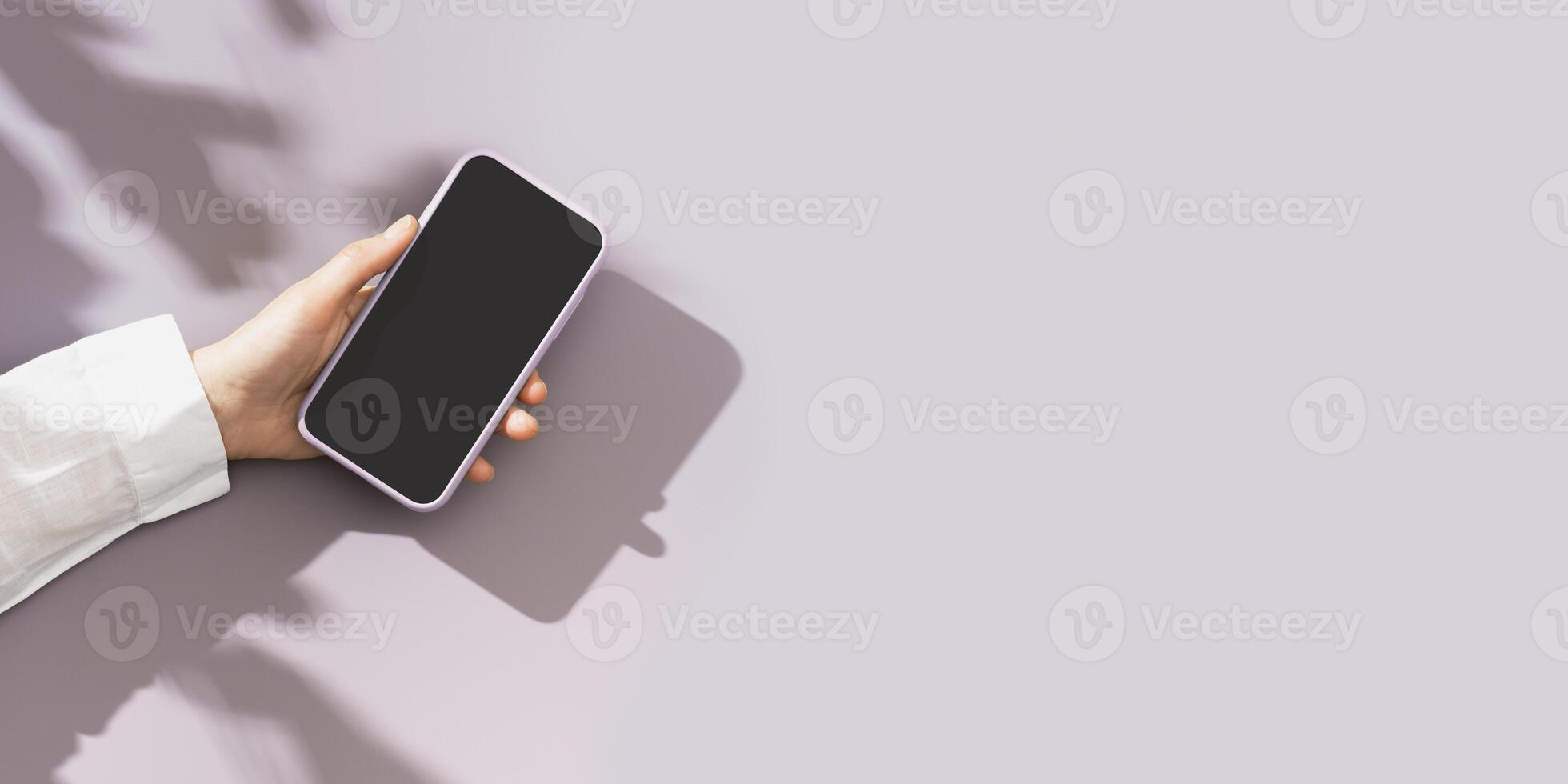 Mock up phone with black blank screen in female hand, white shirt sleeve, minimal flat lay on pastel violet background. Summer lifestyle scene with smartphone, sunlight shadows, wide banner photo