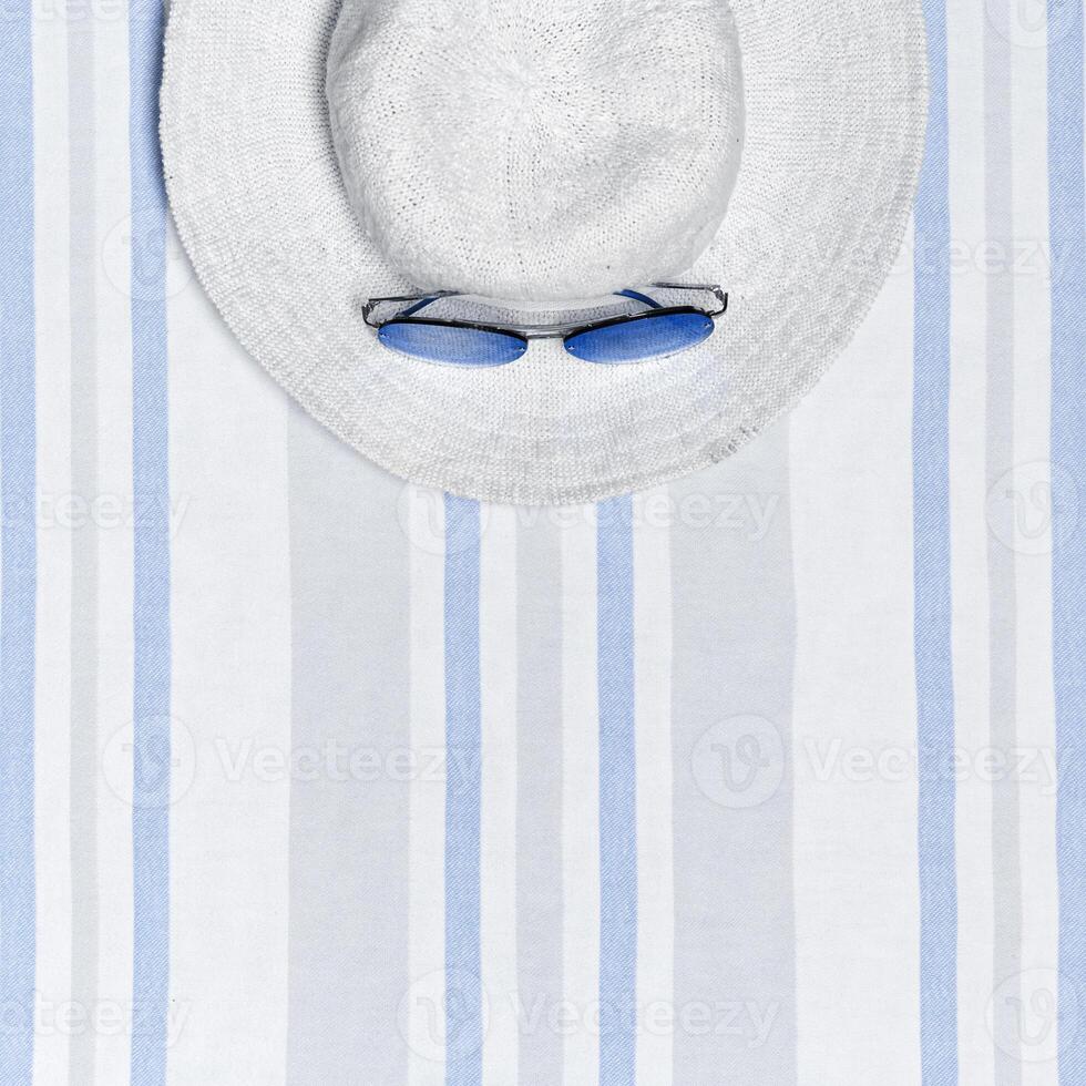 Minimal summer beach concept top view wide-brimmed sun hat and sunglasses on striped beach towel, pastel blue white background. Summertime party, travel, holiday card or invitation. Happy Summer photo