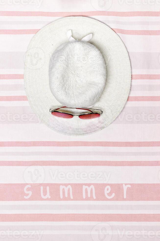 Minimal summer beach concept top view wide-brimmed sun hat with sunglasses on striped beach towel, pastel background. Summertime holiday or vacation card with text summer, pink white flat lay photo
