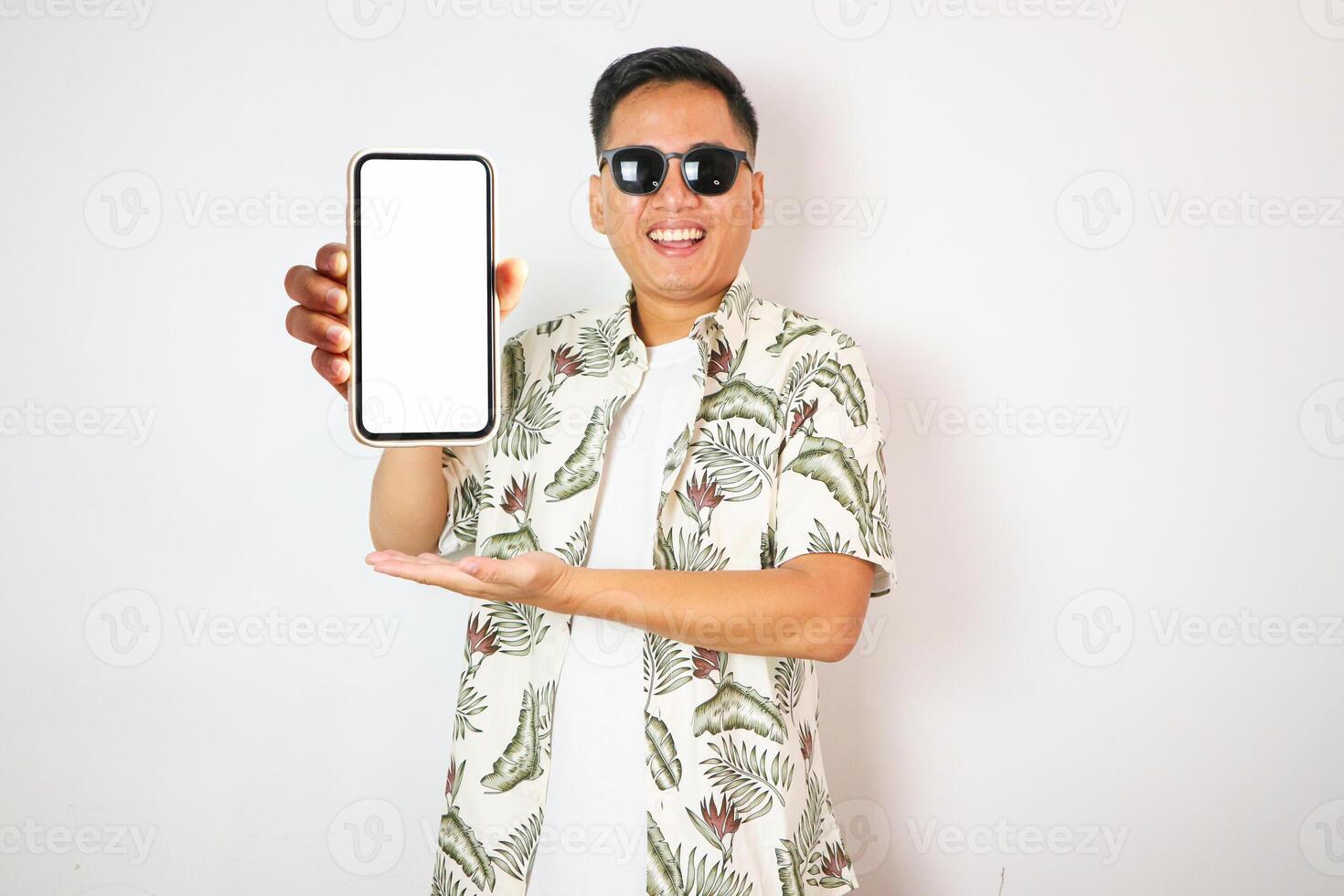 portrait of asian man with gaping or shocked facial expression looking at camera and showing smartphone screen. Indonesian man in Hawaiian shirt on white background isolated. photo