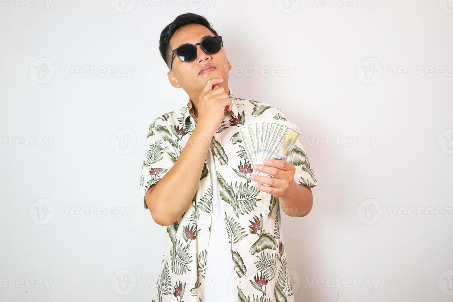 portrait of smiling asian man holding and showing money. portrait of indonesian man in hawaiian shirt looking at camera on isolated white background. gesture showing or thinking photo