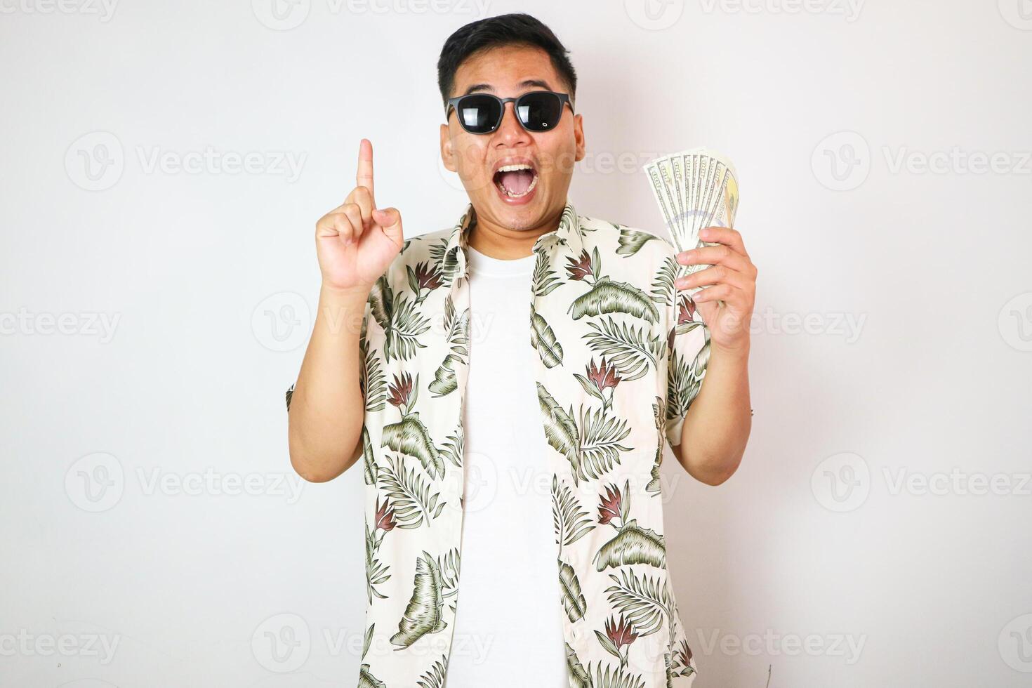 asian man wearing summer and hawaiian lei holding bunch of dollars pointing and showing with thumb up to the side with happy face smiling. photo