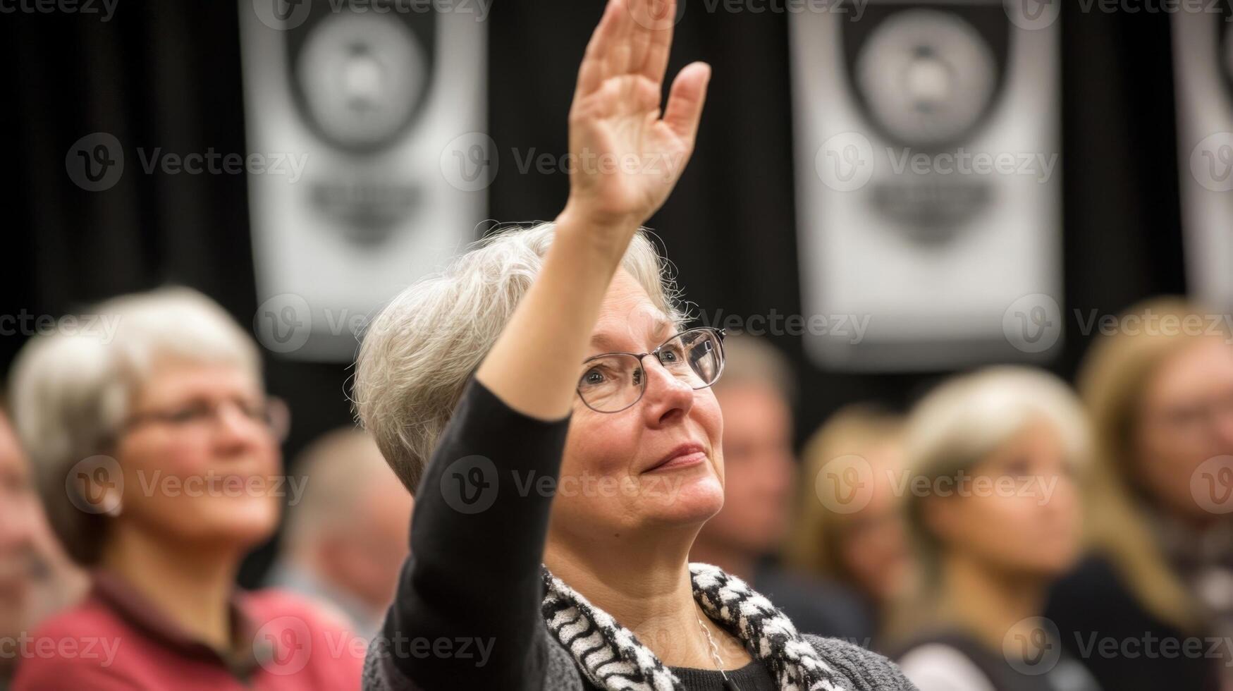 A medium closeup captures a middleaged woman in the audience raising her hand with an expression of curiosity. The backdrop features banners with the town halls logo while other attendee photo