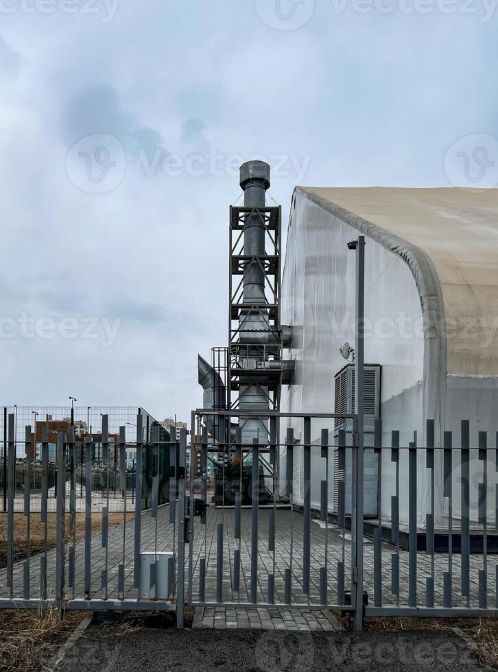 Industrial building with metal chimney and fenced perimeter, showcasing side view of rectangular structure with grey roofing and urban landscape in the background, under cloudy skies photo