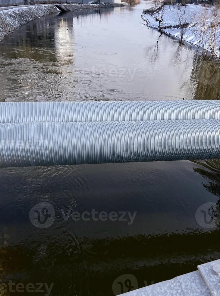 Industrial pipes span across a river in winter, with snow-dusted banks and urban structures, serene waterway photo