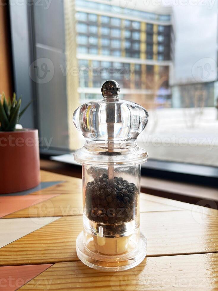 Transparent pepper grinder filled with black peppercorns placed on a wooden table. Modern urban view through a large window in the background. Casual dining setting photo