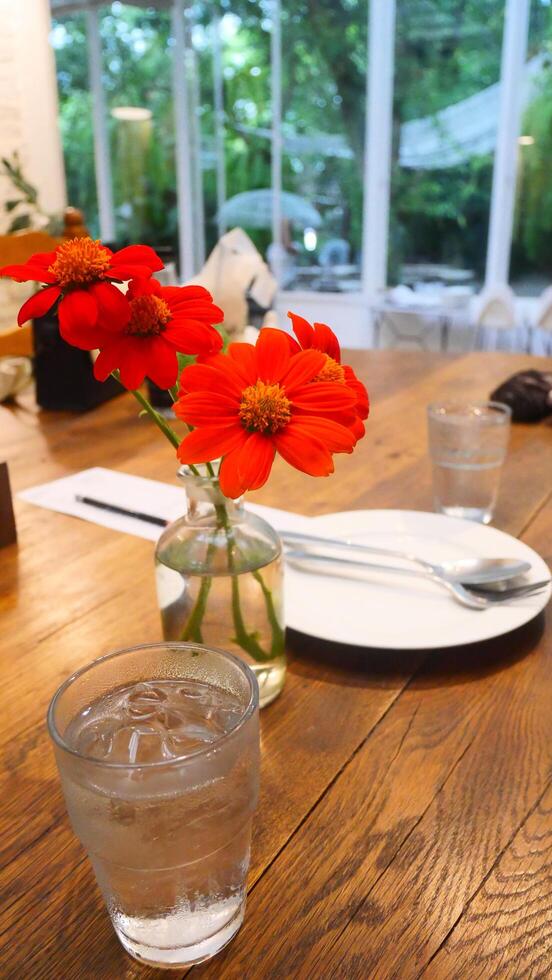 A flower vase, glass of water, plate, spoon, and fork are beautifully arranged on a wooden table in a cafe photo