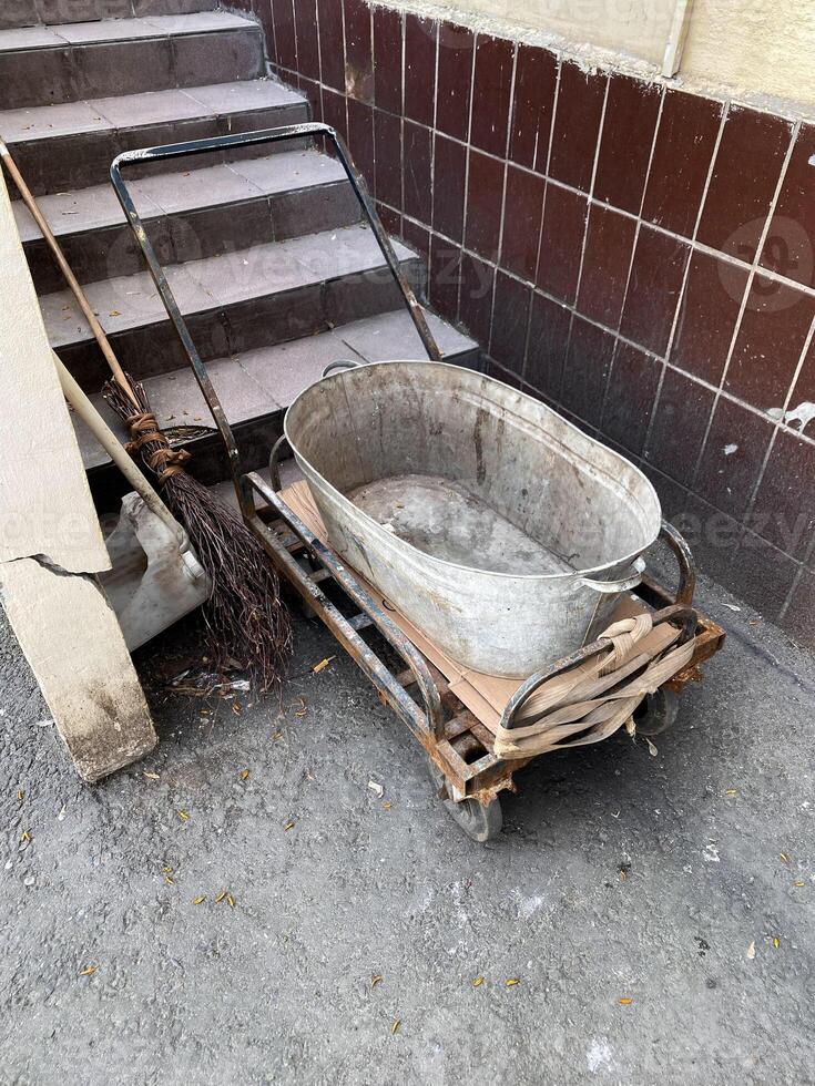 Weathered metal tube on wooden cart, resting on gravel surface next to set of tiled stairs. Signs of wear and broom leans against wall. Typical back alley in residential neighborhood, everyday life photo
