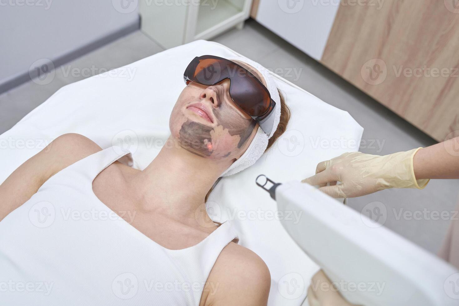 Female caucasian adult undergoing facial laser treatment in a spa clinic setting with protective eye gear. photo