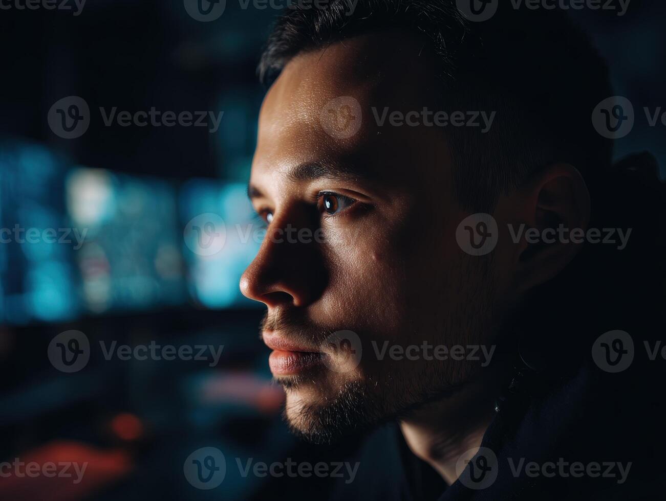 Close-up of a focused individual in dim lighting, gazing intently at multiple computer screens in a dark room, showcasing concentration and a tech-oriented environment photo