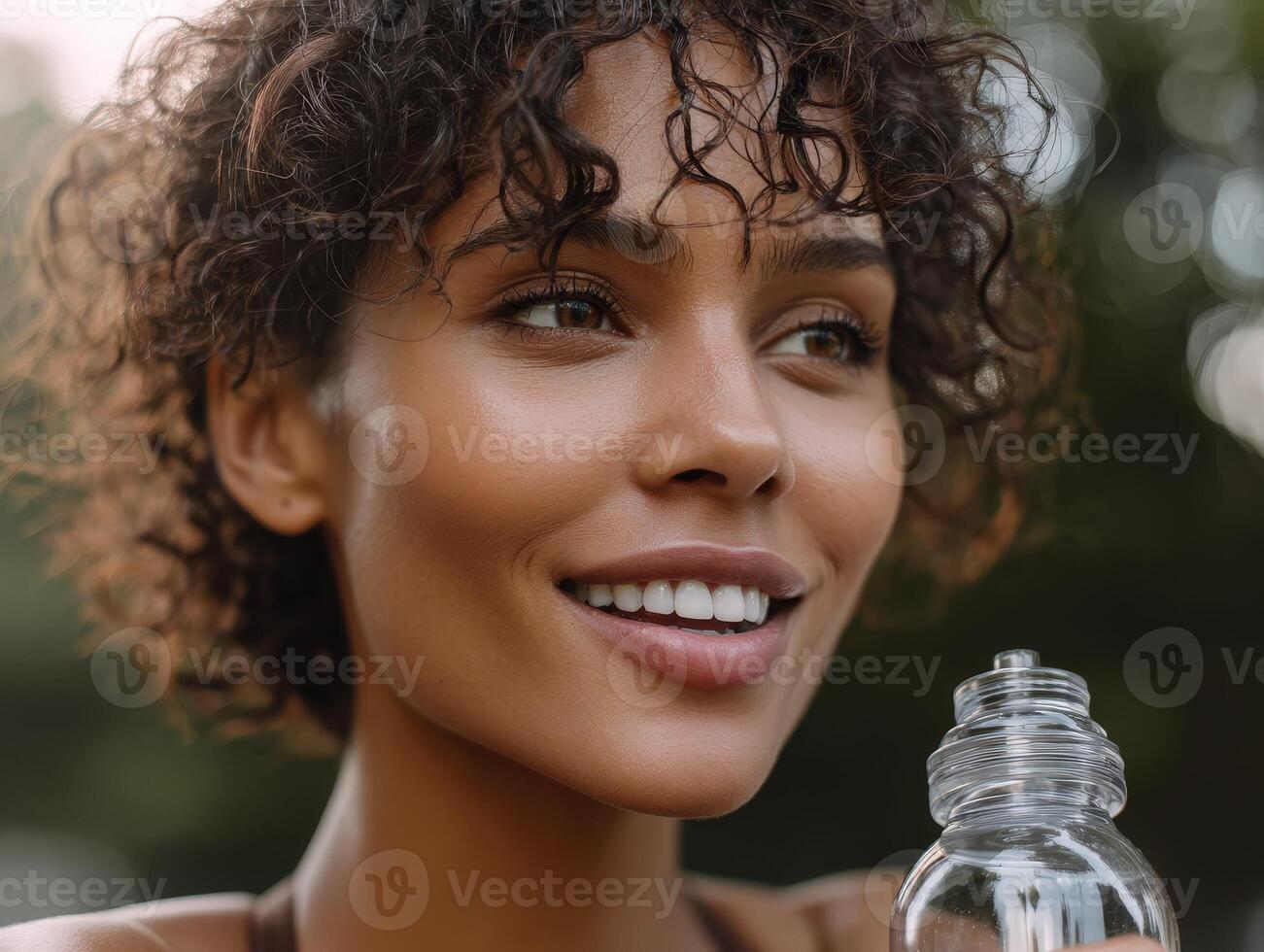 Smiling woman with curly hair holds a clear bottle, showcasing natural beauty and vibrant skin. Close-up portrait, soft bokeh effect in the background highlights her joyful expression photo