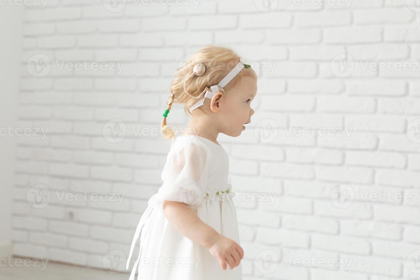 Baby child with hearing aids and cochlear implants and diversity on white brick wall with copy space and place for advertising . Deaf and health concept photo