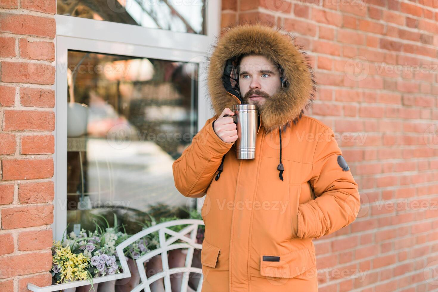 Man in winter coat drinking from thermos on winter city street near shop window. Comfort, routine, and warmth during a cold urban day outdoors. Copy space photo