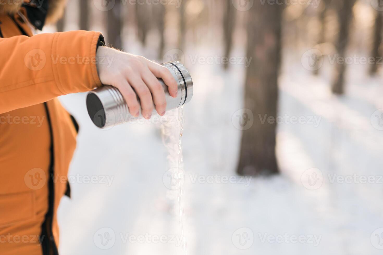 Man pours hot water from a thermos into a snow walking in snowy frozen winter forest at sunset. Adventure, tourism and camping concept. Copy space and empty place for text advertising photo