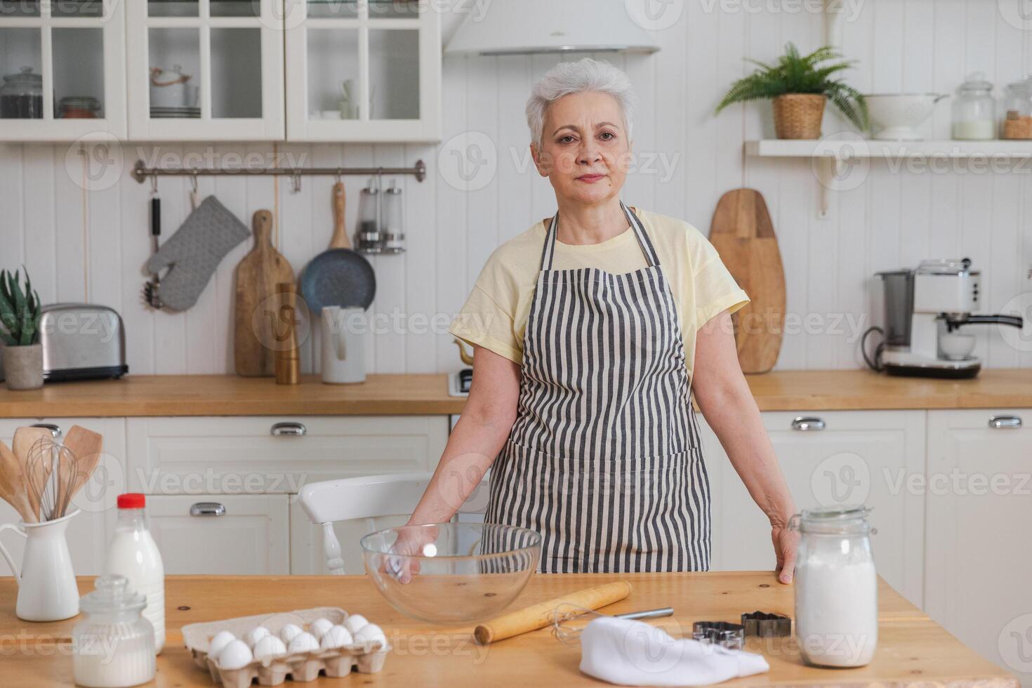 Happy senior woman cooking in kitchen. Stylish older mature gray haired lady grandmother knead dough bake cookies. Old grandma cook homemade food. Household housewife housework concept photo