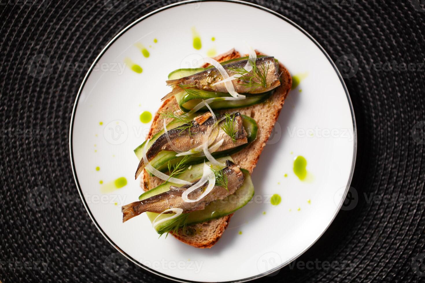 A gourmet open sandwich with sardines on whole grain bread garnished with cucumber slices and green herbs on a white plate photo