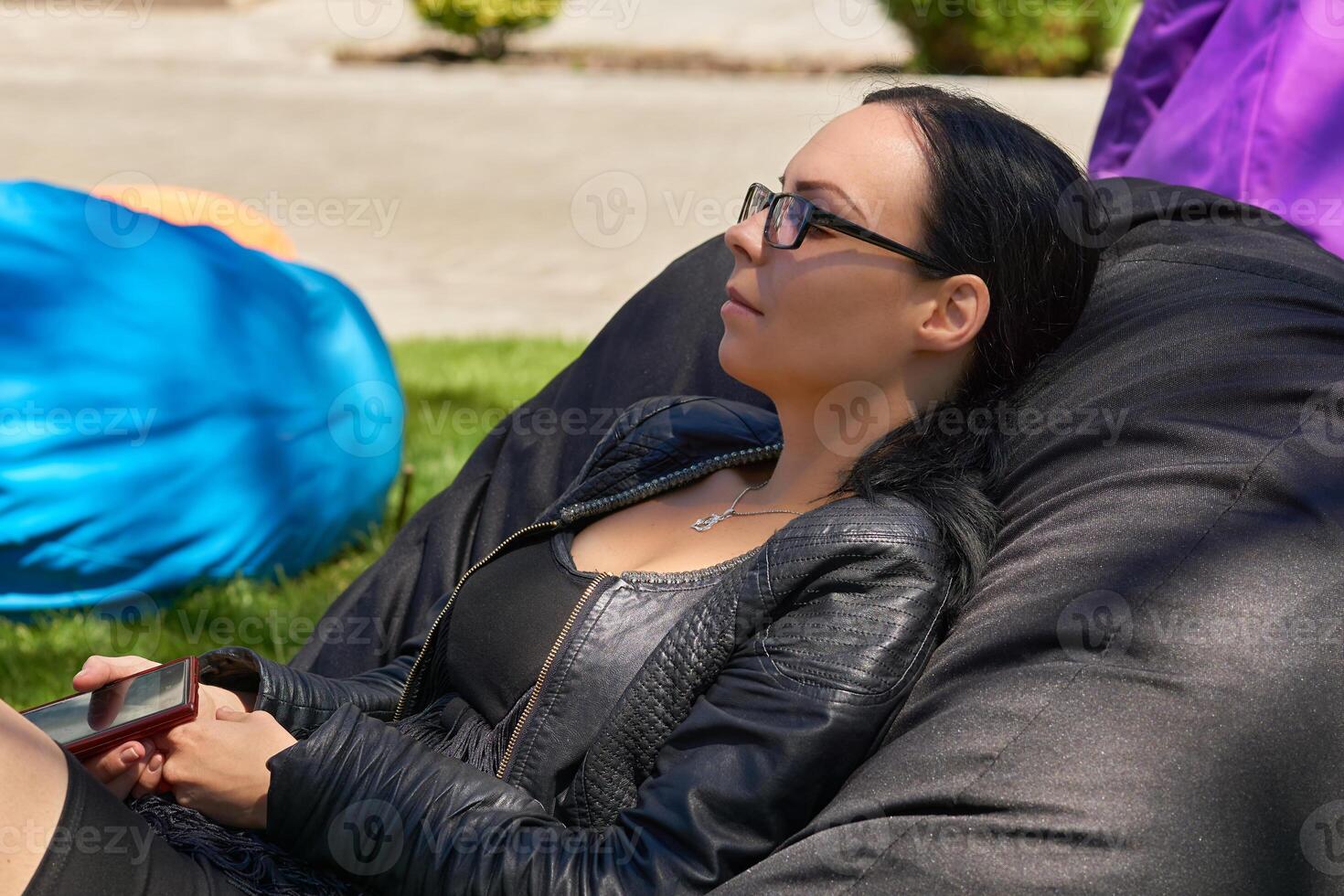 Young woman with glasses resting on a street cushion pouf on a green lawn photo