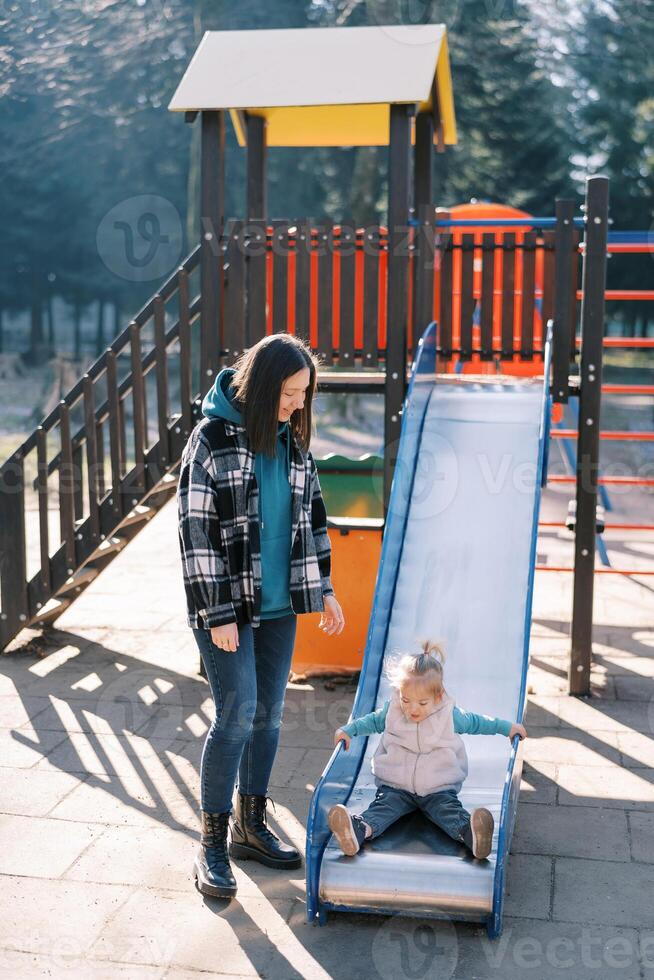 Mom stands next to a little girl sitting on a slide in the playground photo