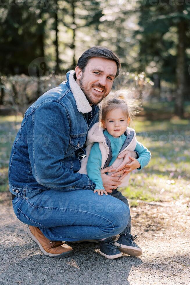 Smiling dad squatting and hugging little daughter in park photo