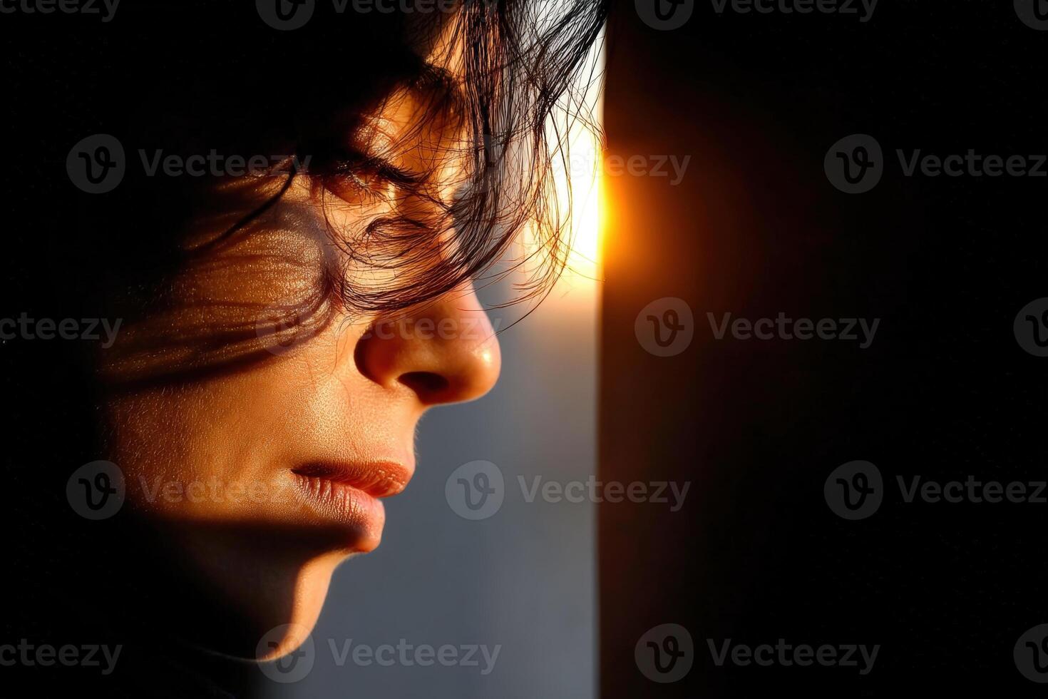 Woman gazing thoughtfully at sunset through a window with soft light illuminating her face photo