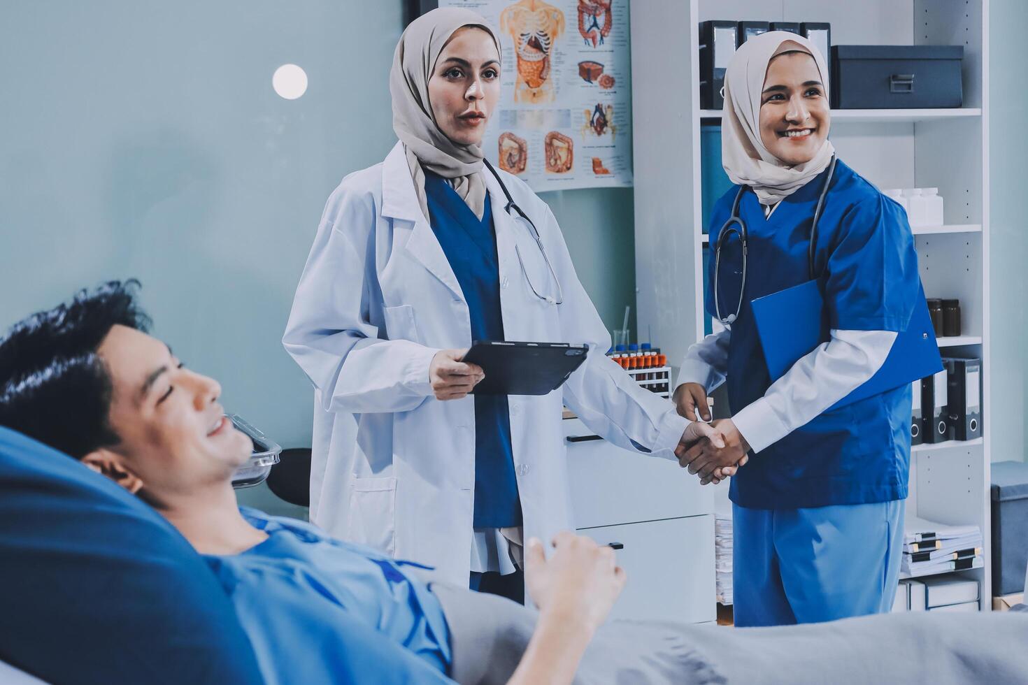 Two women in blue scrubs are shaking hands with a man in a hospital bed photo