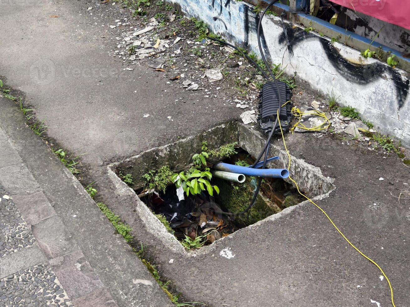 Open sidewalk manhole with exposed cables and dangerous trash-filled hole photo