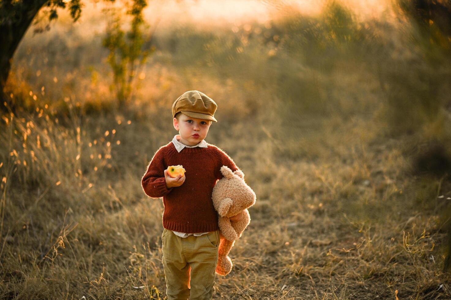 A young boy in vintage clothing walks through a golden field at sunset, holding a big teddy bear and eating apple photo