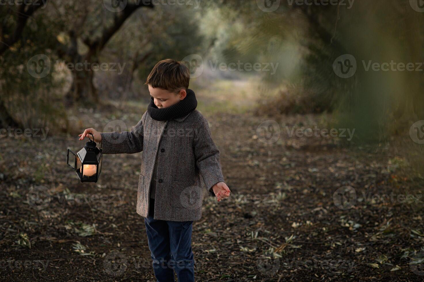 A little boy in a fantasy forest lifts a lantern gently, staring into its glow as the forest around him shimmers with mystery. photo