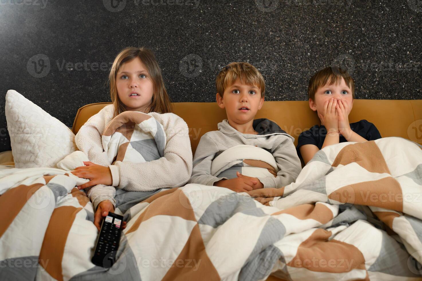 Kids sitting close together on a living room sofa, remote in hand, showing expressions of fear and wonder while watching a suspenseful scene. photo