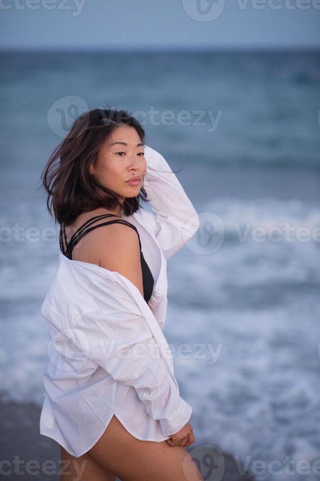 Female in black swimwear and white shirt stands on the coast, eyes closed, enjoying the breeze. photo