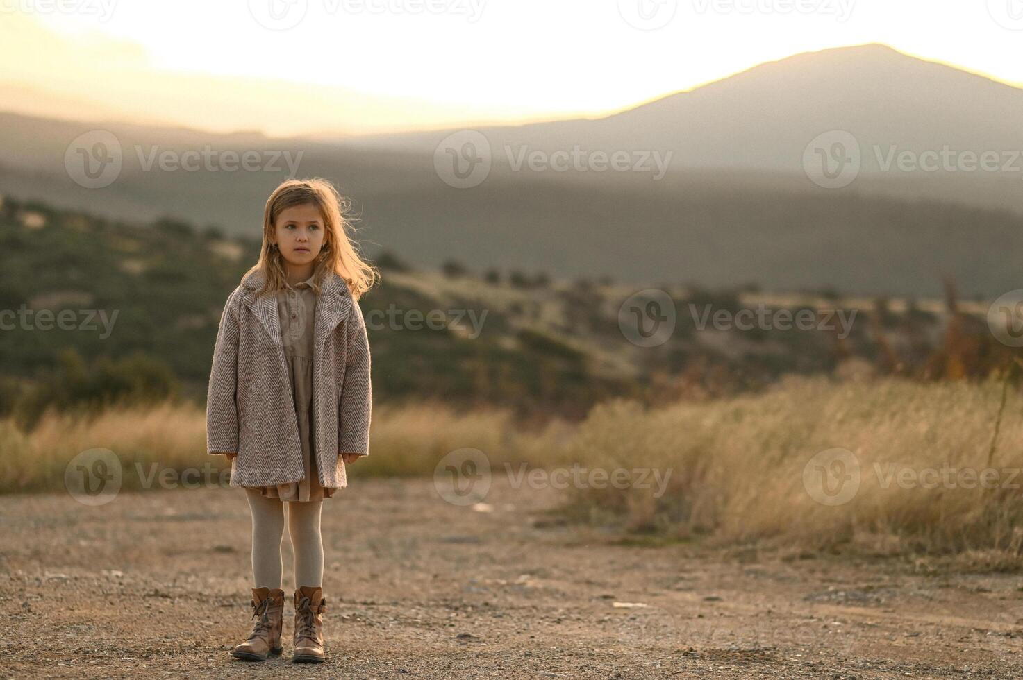 The girl twirls slowly on a sunlit dirt road, her coat flaring out, hair glowing in the warm light. photo