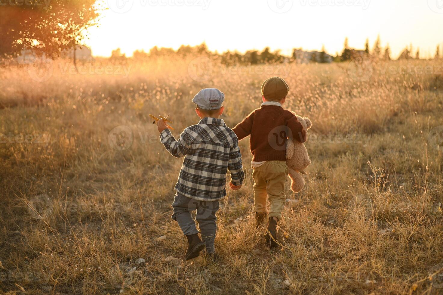 In a warm sunset glow, two young boys in old-fashioned outfits share a quiet moment in a field with an apple and a plush bear. photo