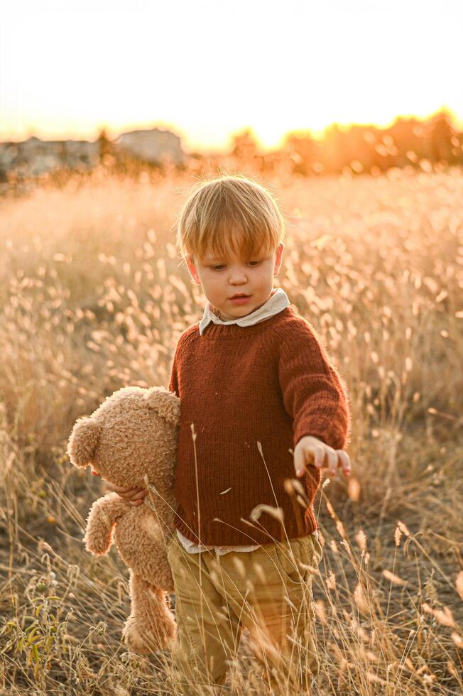 Dressed in old-fashioned clothes, a boy stands in tall grass at golden hour, gazing at the horizon with a large plush bear by his side. photo