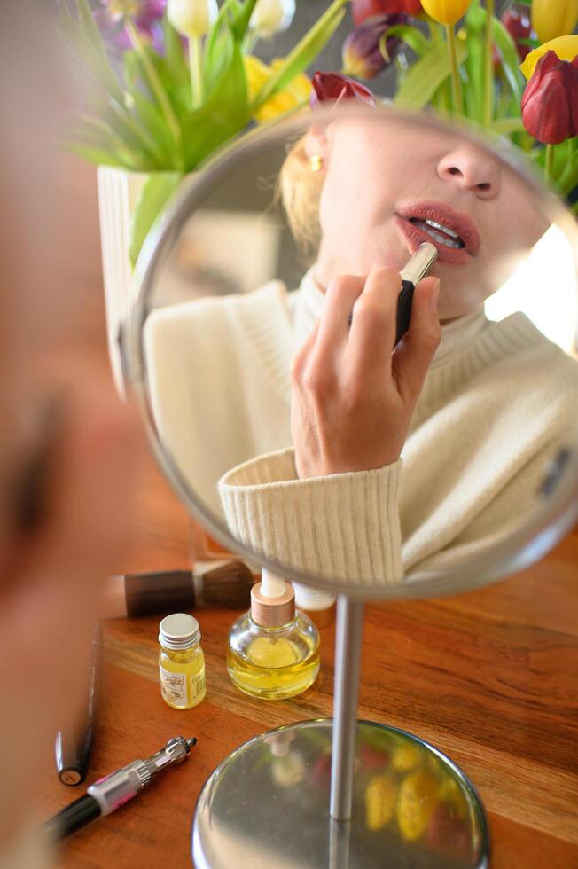 Close-up photo of a woman applying lipstick to her lips in front of a mirror. She has a focused expression as she carefully enhances her makeup, with soft lighting highlighting her features.