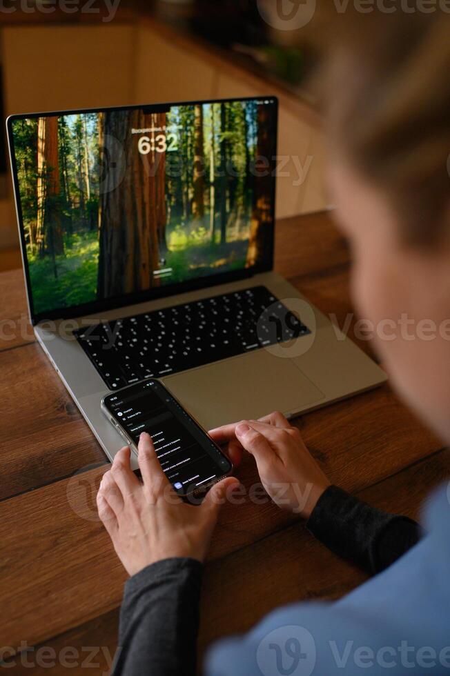 A person sits in front of a laptop, holding a phone showing a chat with ChatGPT on the screen. photo