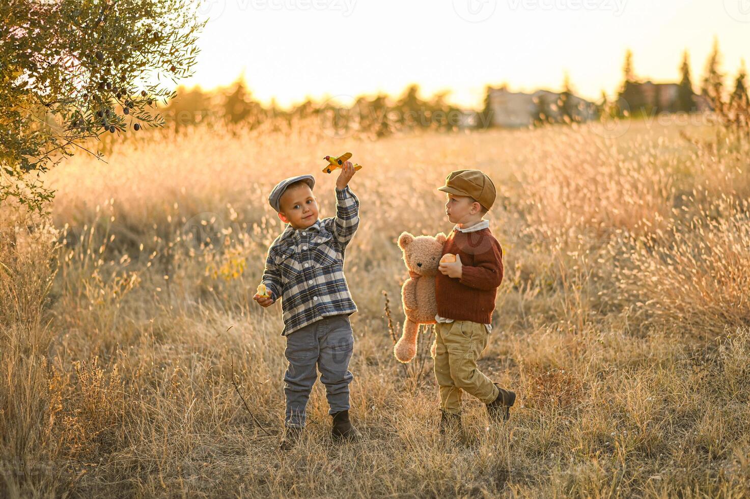 In the warm glow of sunset, two children in old-fashioned outfits laugh and run through a field with an apple, a plush bear, and a toy plane. photo