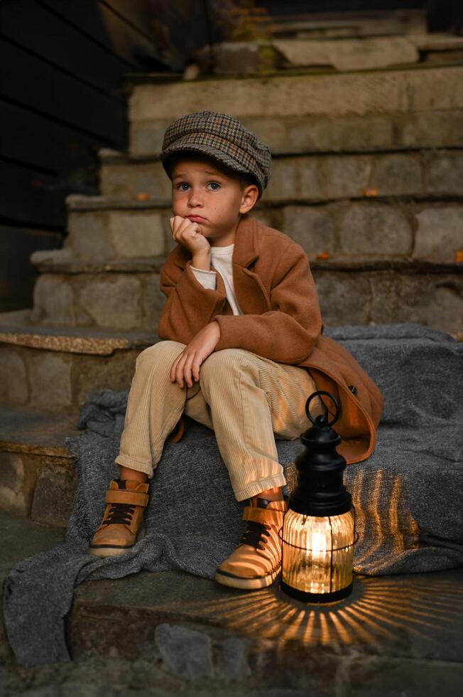 A young boy in vintage clothing sits on the steps beside a glowing lantern, bathed in soft golden light. photo