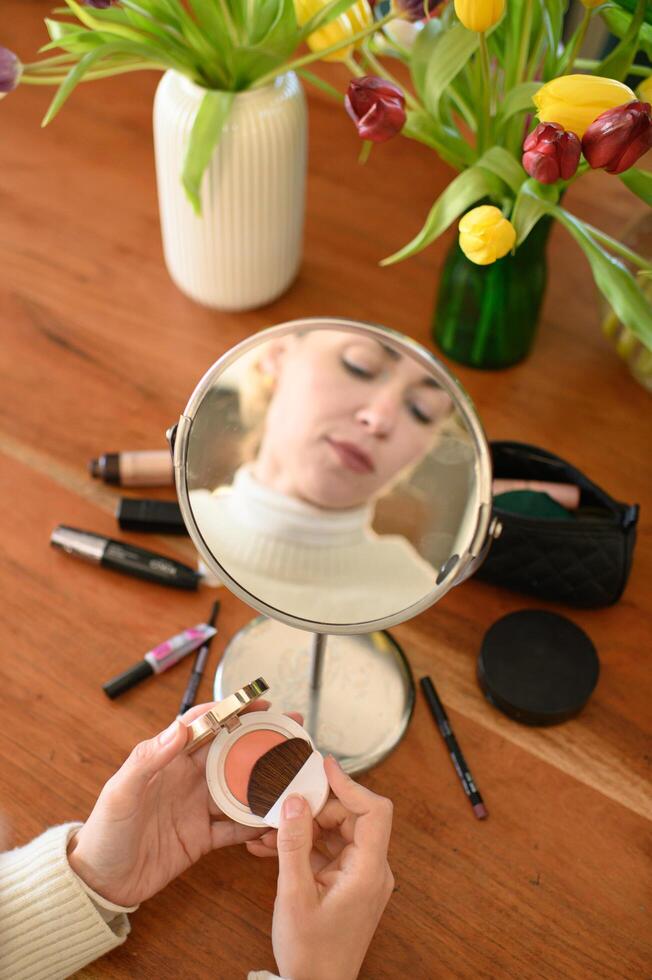 A woman sits in front of a mirror holding a blush compact, with her reflection visible as she prepares to apply it. photo