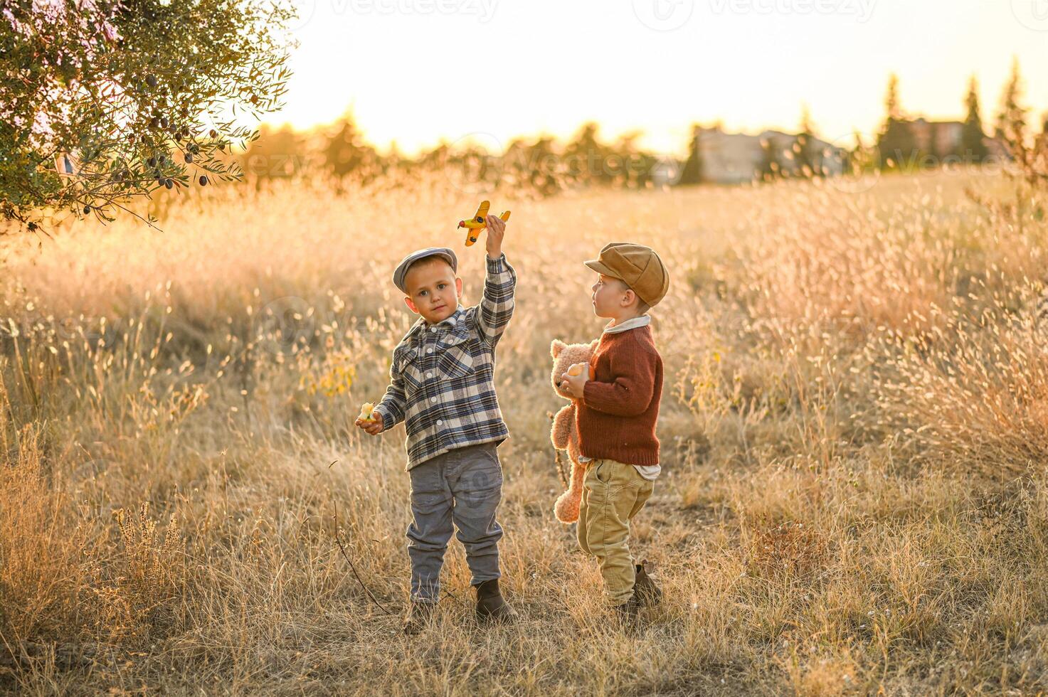 Bathed in golden light, two boys in classic vintage outfits play joyfully with a teddy bear, a small airplane, and a shiny apple in hand. photo