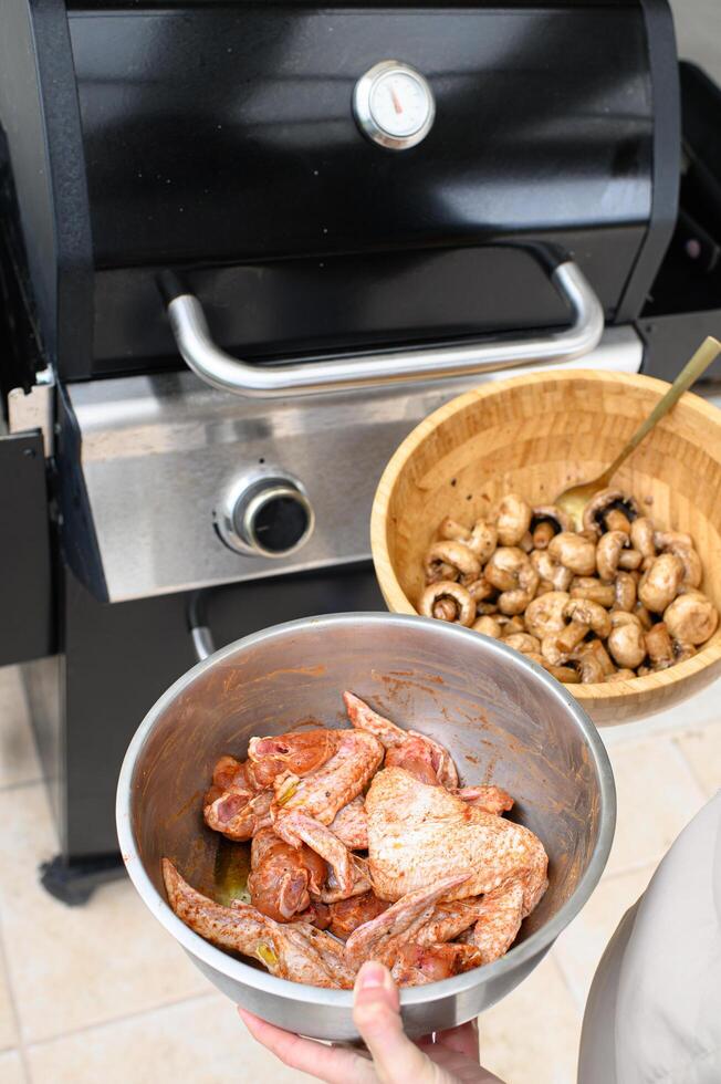 A pair of hands holds a platter with marinated chicken and mushrooms over a lit grill. photo