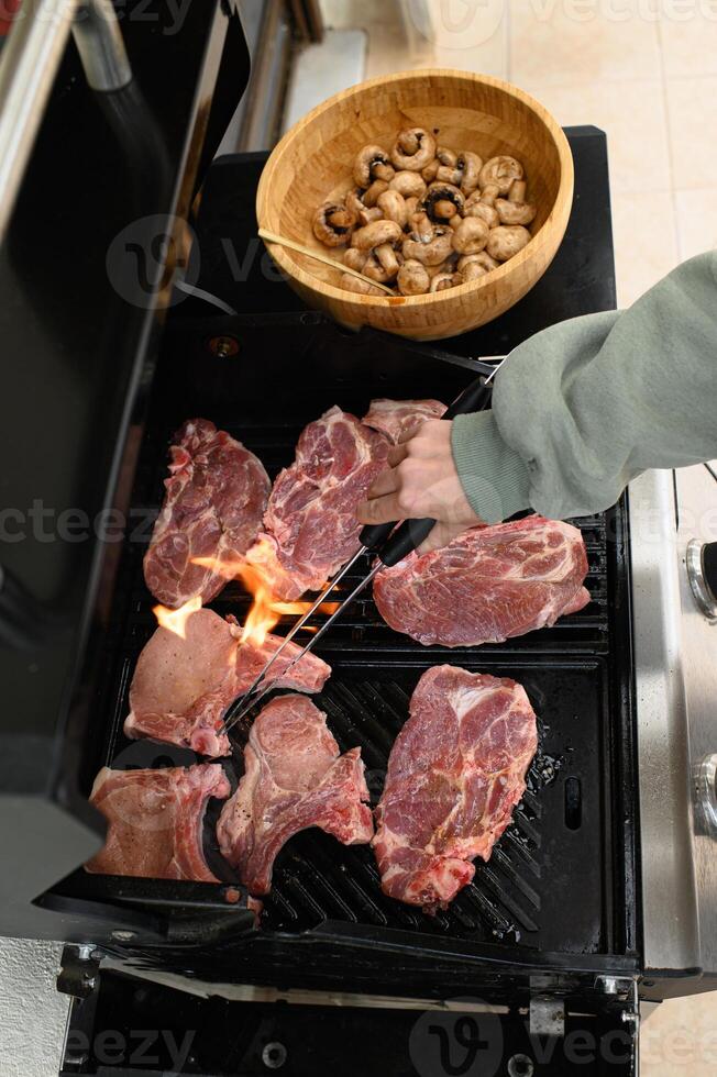 One hand places meat on the grill with tongs, the other steadying a plate of mushroom skewers. photo