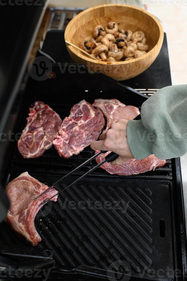 Close-up of hands using tongs to lay fresh meat slices onto the barbecue, mushrooms waiting on a dish beside. photo