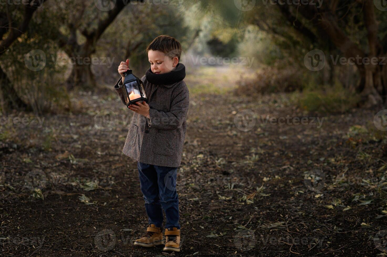 A small boy pauses under tall glowing trees, mesmerized by the golden light of his lantern in the quiet forest. photo