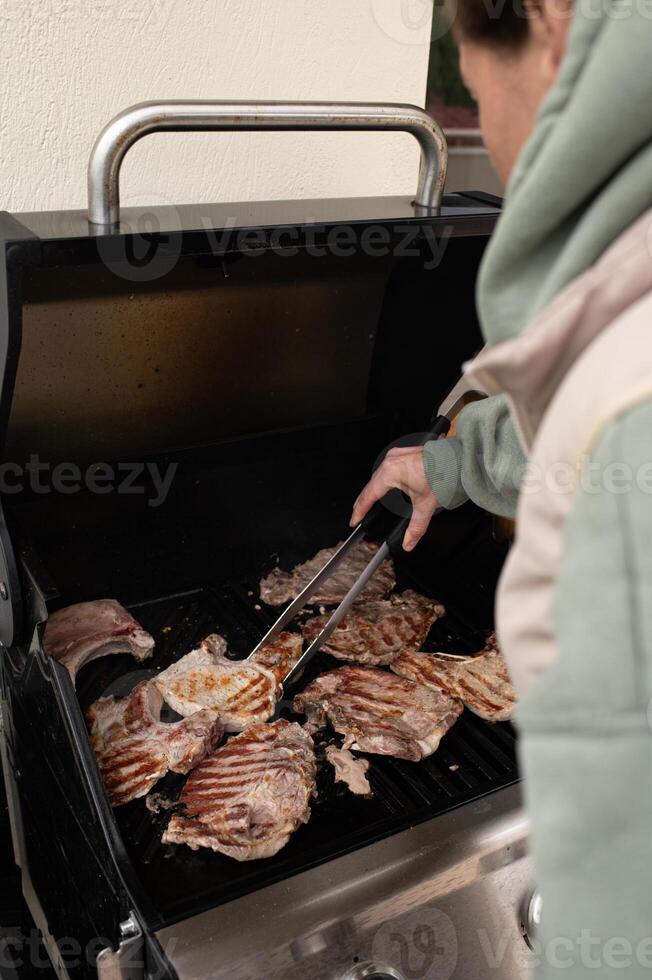 Over a fiery barbecue, a man in a casual shirt tends to the meat, using tongs to flip a thick piece of steak as smoke swirls. photo