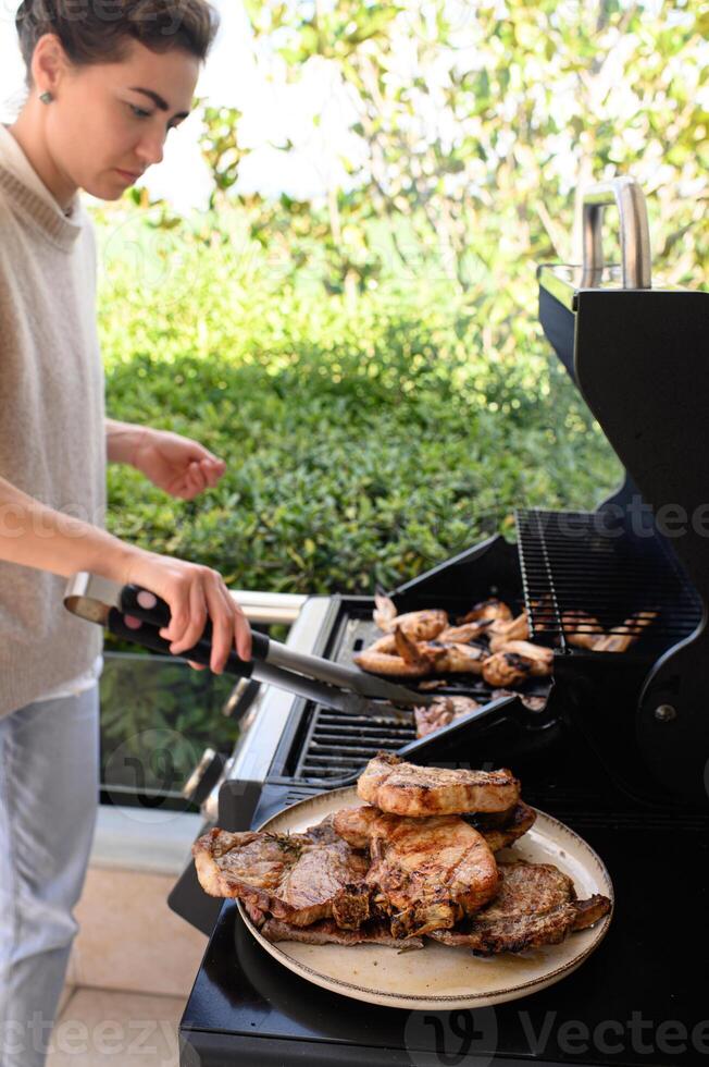 With a smile, a woman balances the grill with one hand and serves fresh grilled meat and chicken onto a plate with the other, enjoying the outdoor cookout. photo