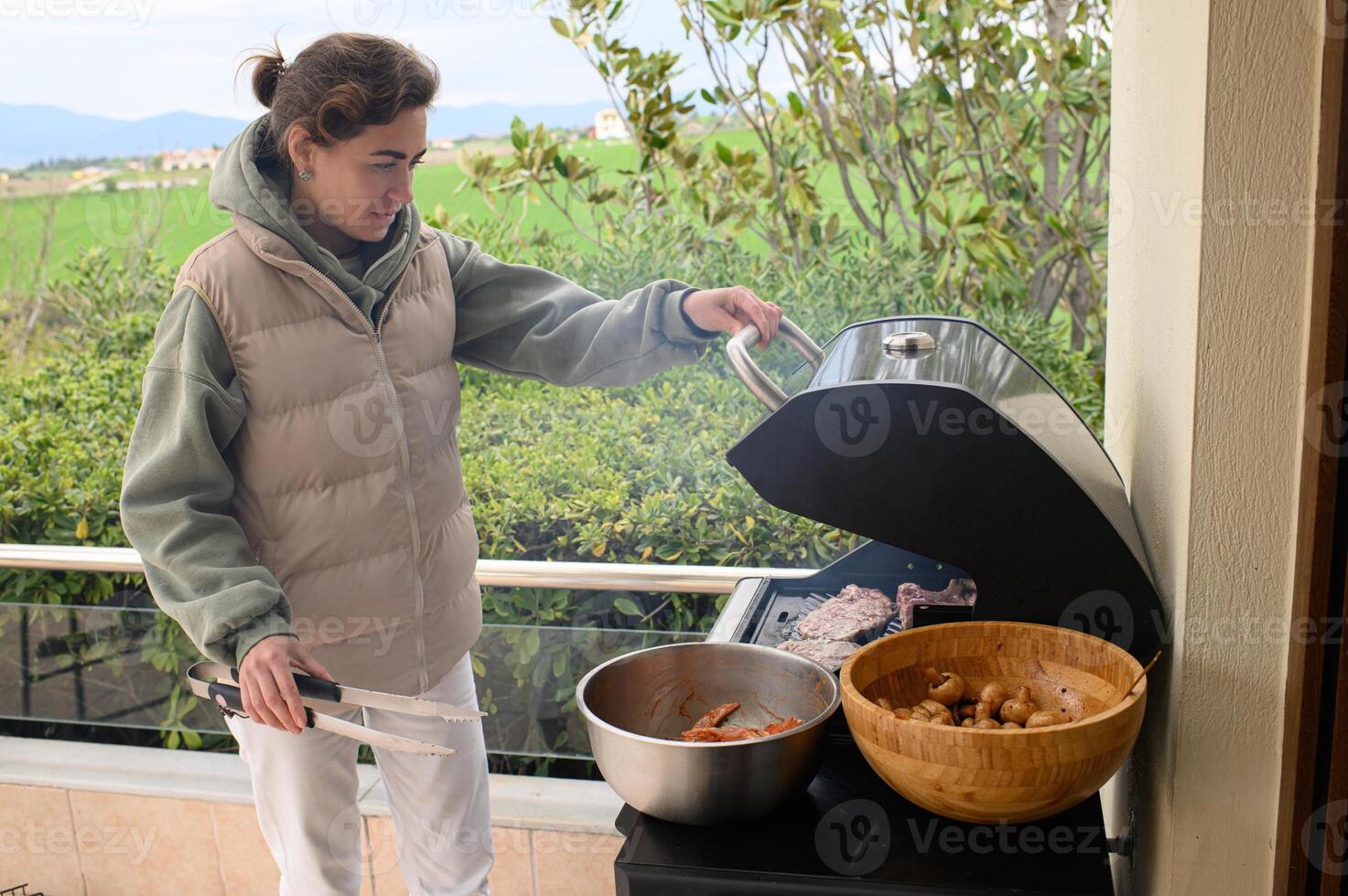 A relaxed young woman by the grill on a rooftop terrace prepares to cook, with meat already searing and more chicken and mushrooms waiting. photo