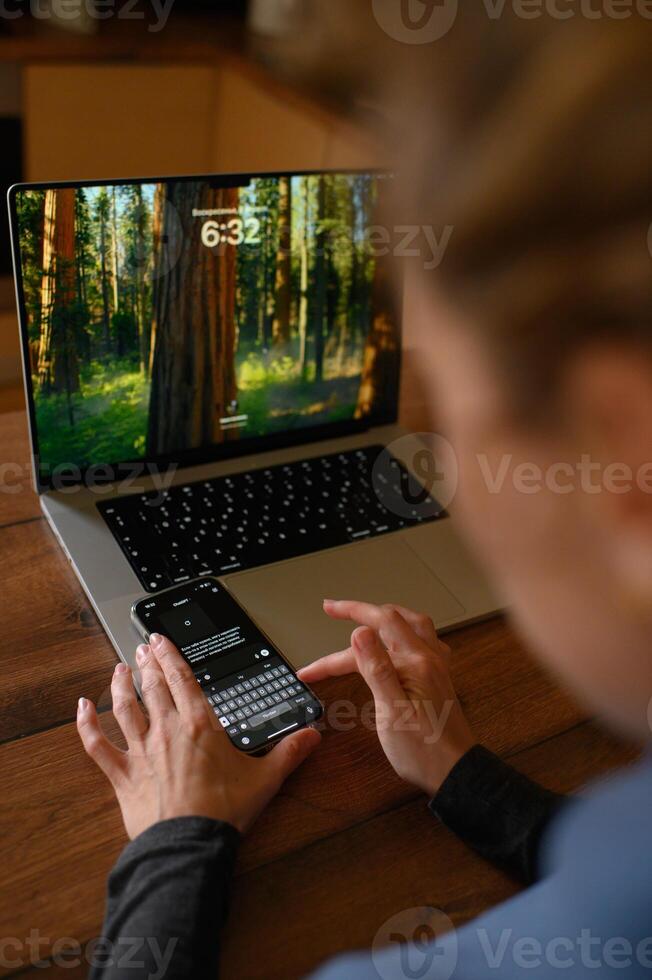 Someone holds a phone with an active ChatGPT conversation, seated at a workspace with a laptop in front. photo