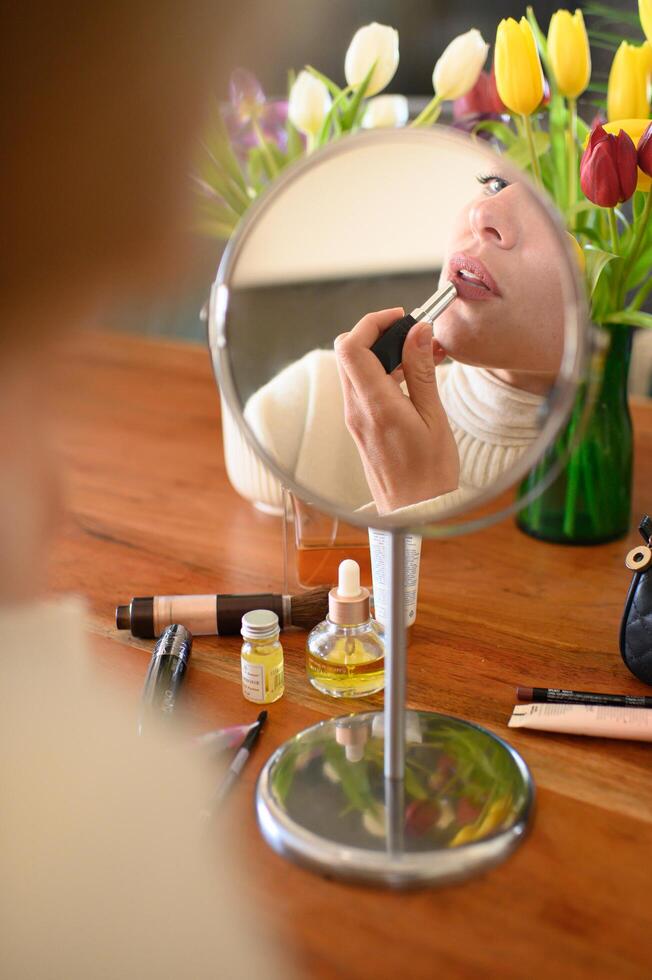 A close up of a womans lips reflected in a mirror as she applies lipstick, with scattered makeup products on the table. photo